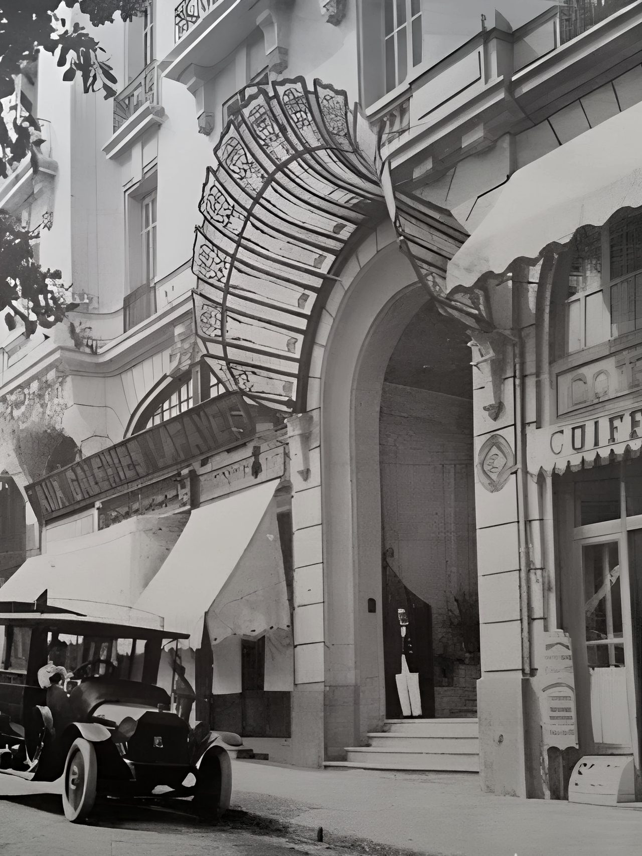 Photographie de l’arcade d’entrée, surmontée de la marquise en éventail de fer et de verre. Une automobile des années 1920 est arrêtée devant le perron ; à droite, l’enseigne d’un coiffeur.